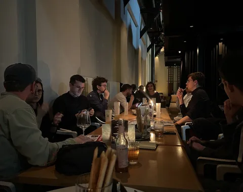 Alumni-Treffen in einem gemütlichen Restaurant: Eine Gruppe von Menschen sitzt an einem langen Tisch, unterhält sich und genießt Getränke in entspannter Atmosphäre.