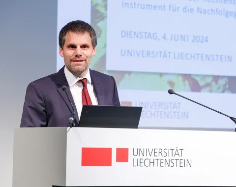Ein Mann in einem Anzug steht an einem Rednerpult mit dem Logo der Universität Liechtenstein. Im Hintergrund ist eine Präsentationsfolie mit dem Datum 4. Juni 2024 zu sehen.