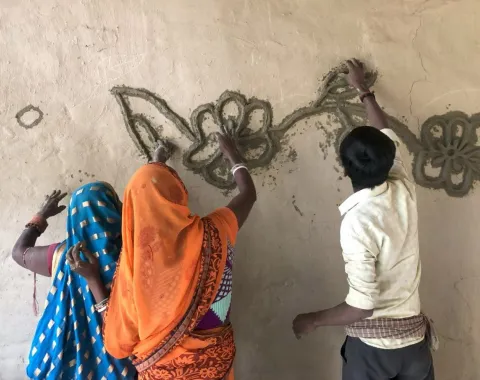 Drei Personen gestalten gemeinsam eine Wand mit traditionellen Mustern aus Lehm. Sie tragen bunte Kleidung und arbeiten konzentriert an einem floralen Design.