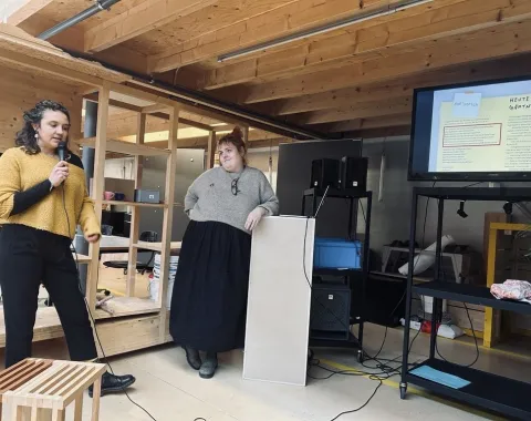 Zwei Personen präsentieren in einem Raum mit Holzelementen. Eine Person spricht in ein Mikrofon, die andere steht neben einem Rednerpult. Im Hintergrund ist eine Leinwand mit einer Präsentation zu sehen.