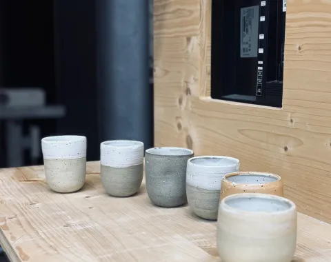 Sechs handgetöpferte Keramiktassen in verschiedenen Erdtönen stehen auf einem Holztisch. Die Tassen sind schlicht und modern gestaltet, passend zur Architekturveranstaltung an der Universität Liechtenstein.