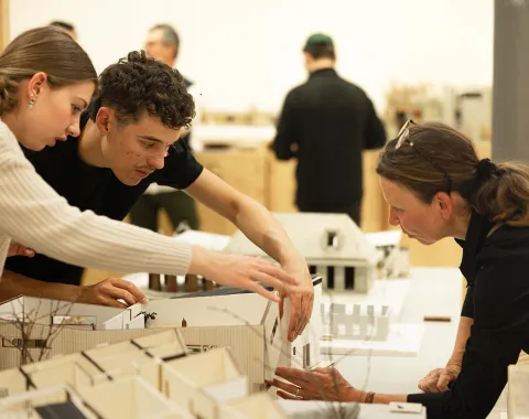 Drei Personen betrachten und diskutieren über Architekturmodelle auf einem Tisch in einem Ausstellungsraum. Im Hintergrund sind weitere Personen und Modelle zu sehen.