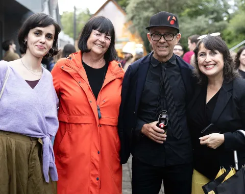 Vier Personen stehen lächelnd nebeneinander bei einer Veranstaltung im Freien. Sie tragen unterschiedliche, farbenfrohe Kleidung. Im Hintergrund sind weitere Menschen und Bäume zu sehen.