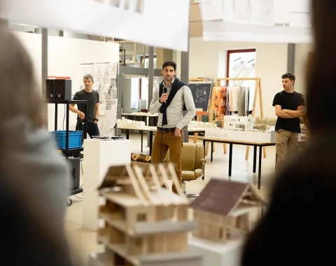 Ein Redner hält einen Vortrag in einem Ausstellungsraum der Universität Liechtenstein. Im Vordergrund sind Architekturmodelle zu sehen. Zuschauer hören aufmerksam zu.