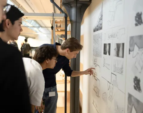 Drei Personen betrachten und diskutieren Architekturzeichnungen an einer Wand in einem modernen Innenraum. Eine Person zeigt auf Details der Zeichnungen. Die Szene vermittelt eine Atmosphäre des Lernens und der Zusammenarbeit.