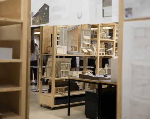 Architekturmodelle und Zeichnungen in einem Ausstellungsraum der Universität Liechtenstein. Holzregale präsentieren detaillierte Miniaturgebäude und Baupläne. Im Hintergrund sind Personen unscharf zu erkennen.