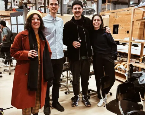 Vier Personen stehen lächelnd in einem Architekturstudio. Sie tragen lässige Kleidung und halten Getränke in der Hand. Im Hintergrund sind Arbeitsplätze und Modelle zu sehen. Die Atmosphäre wirkt freundlich und kreativ.