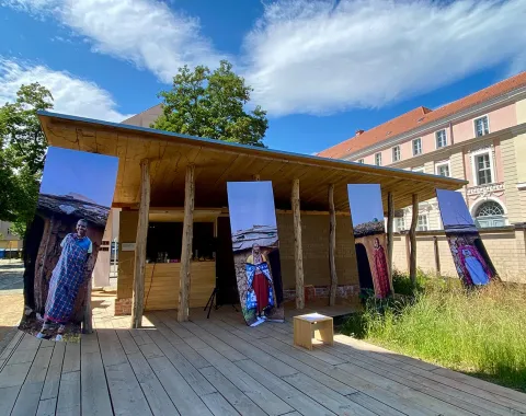 Ein Holzpavillon mit großen, vertikalen Fotobannern, die Maasai-Baumeisterinnen zeigen, steht auf einer Holzplattform. Im Hintergrund sind historische Gebäude und ein blauer Himmel mit Wolken zu sehen. Die Szene ist Teil einer Ausstellung über weibliche Perspektiven in der Architektur.