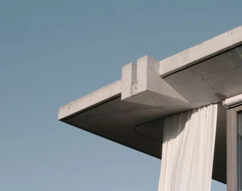 Betonvordach mit klaren Linien und hängendem weißen Vorhang vor blauem Himmel. Moderne Architektur, die Minimalismus und Eleganz vereint.