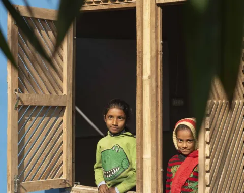 Zwei Kinder schauen aus einem offenen Fenster eines Gebäudes mit blauer Wand und dekorativem Muster. Das Fenster hat hölzerne Fensterläden. Im Vordergrund sind unscharfe Blätter zu sehen.