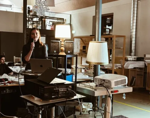 Eine Person hält eine Präsentation in einem modernen Raum mit technischen Geräten und Lampen. Im Hintergrund sind Regale und Arbeitsmaterialien zu sehen.