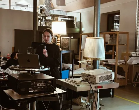 Eine Person hält eine Präsentation in einem modernen Raum mit technischer Ausstattung. Im Vordergrund stehen ein Laptop und ein Projektor auf einem Tisch. Der Raum ist gut beleuchtet und im Hintergrund sind Regale und weitere Personen zu sehen.