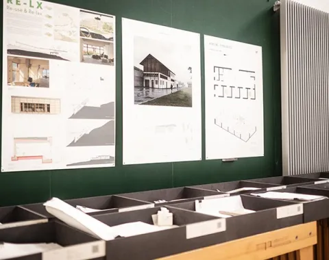 Architektur-Entwürfe und Pläne an einer Wand in der Liechtenstein School of Architecture. Modelle und Zeichnungen sind in Regalen darunter angeordnet.