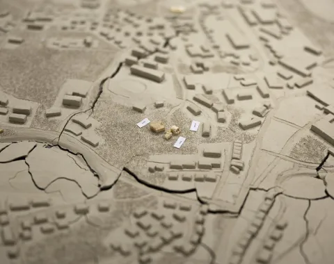 Ein detailliertes Architekturmodell zeigt eine Stadtlandschaft mit Gebäuden und Straßen. Im Zentrum sind mehrere kleine, beschriftete Modellelemente platziert, die möglicherweise auf ein spezifisches Bauprojekt hinweisen. Das Modell bietet eine Vogelperspektive auf die städtische Umgebung und dient als visuelles Hilfsmittel für architektonische Planungen und Präsentationen.