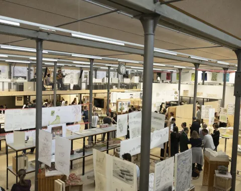 Innenansicht eines belebten Architekturstudios mit zahlreichen Menschen, die sich unterhalten und Arbeiten betrachten. Zeichnungen und Pläne hängen von der Decke. Die Szene vermittelt eine kreative und kollaborative Atmosphäre.