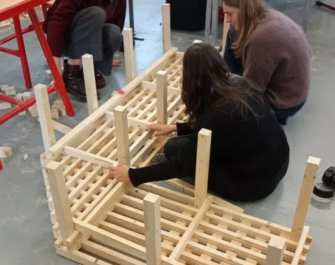 Drei Studierende arbeiten gemeinsam an einem Holzprojekt in einer Werkstatt. Sie montieren ein Möbelstück aus Holzlatten. Im Hintergrund sind Werkbänke und Werkzeuge sichtbar.