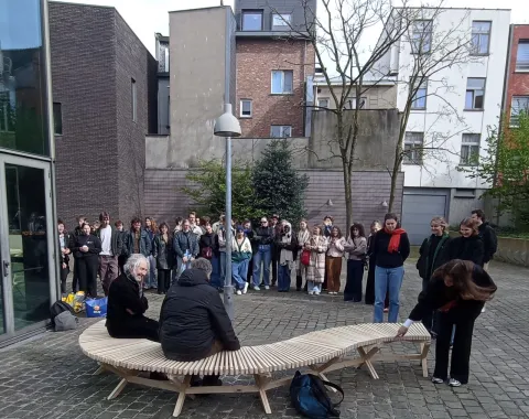 Eine Gruppe von Studierenden steht im Freien um eine geschwungene Holzbank, die von zwei Personen genutzt wird. Die Szene findet auf einem gepflasterten Platz vor modernen Gebäuden statt.