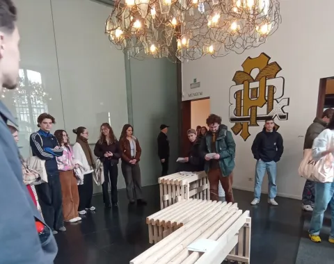 Eine Gruppe von Studierenden steht in einem Raum der Universität Antwerpen um ein modernes, aus Holzlatten gefertigtes Möbelstück. Über ihnen hängt eine große, kunstvolle Lampe. Im Hintergrund ist ein Wandlogo der Universität zu sehen.