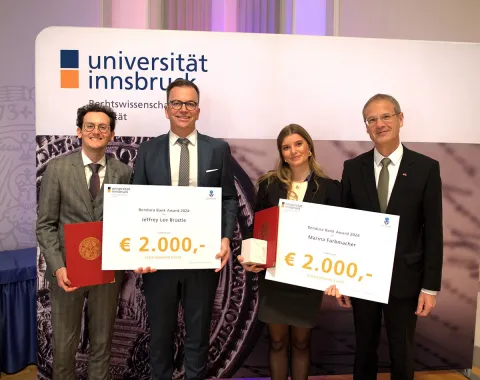 Vier Personen stehen nebeneinander und halten große Schecks mit der Aufschrift "Bendura Bank Award 2024" und einem Betrag von 2.000 Euro. Im Hintergrund ist ein Banner der Universität Innsbruck zu sehen. Die Personen sind formell gekleidet und lächeln in die Kamera.