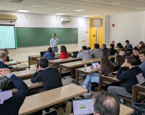 Ein Dozent hält einen Vortrag in einem Hörsaal, umgeben von Studierenden, die aufmerksam zuhören und Notizen auf Laptops machen. Im Hintergrund ist eine Tafel und eine Präsentation zu sehen.