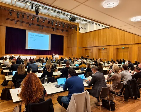 Ein großer Konferenzraum mit vielen Teilnehmern, die an Tischen sitzen und einem Vortrag folgen. Eine Präsentation wird auf einer Leinwand gezeigt. Die Veranstaltung findet in einem modernen Saal mit Holzwänden statt.