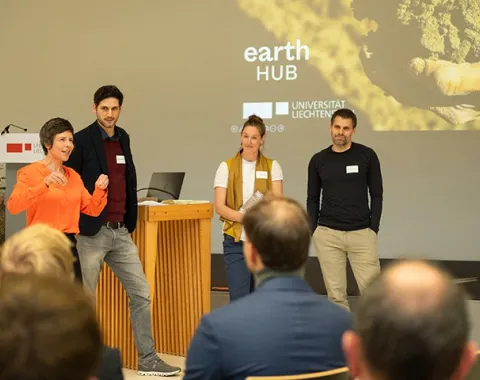 Eine Gruppe von vier Personen steht vor einem Publikum und spricht auf einer Bühne. Im Hintergrund ist eine Präsentation mit dem Titel "Earth Hub" und dem Logo der Universität Liechtenstein zu sehen. Die Szene findet in einem Konferenzraum statt.