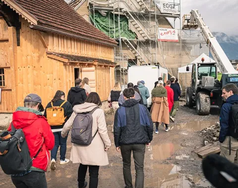 Eine Gruppe von Menschen besichtigt eine Baustelle mit einem Holzgebäude und Baugerüsten. Im Hintergrund sind Berge zu sehen. Die Szene wirkt lebendig und zeigt das Interesse an nachhaltigem Bauen.