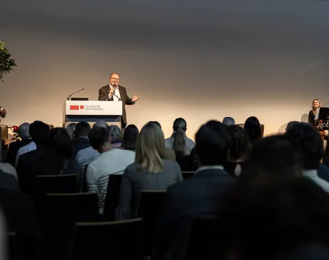Ein Redner steht an einem Podium mit dem Logo der Universität Liechtenstein, spricht zu einem Publikum. Im Hintergrund sind zwei Musiker zu sehen. Die Veranstaltung findet in einem großen Saal statt.