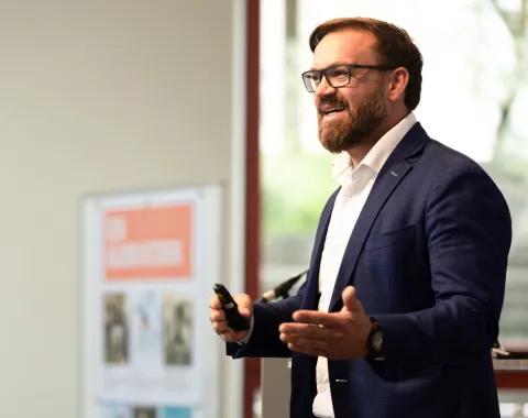 Auf dem Bild ist der Studiengangsmanager des Masters in Entrepreneurship & Management Tobias Fitz zu sehen wie er gerade eine Rede hält. Er trägt einen blauen Anzug mit einem weissen Hemd.