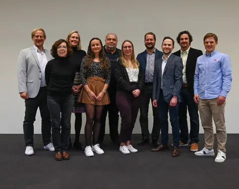 Eine Gruppe von zehn Personen steht lächelnd nebeneinander vor einem neutralen Hintergrund. Sie tragen unterschiedliche, teils formelle, teils legere Kleidung. Das Bild vermittelt eine freundliche und professionelle Atmosphäre.