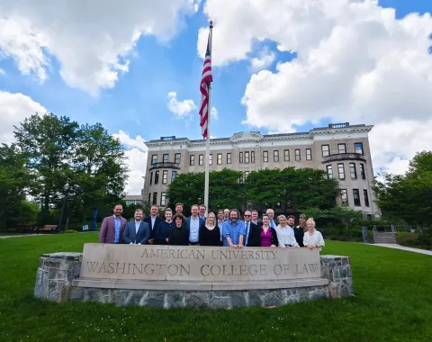 Gruppe von Studierenden und Dozenten des Executive Master of Laws der Universität Liechtenstein vor dem Washington College of Law der American University, mit einer US-Flagge im Vordergrund und einem historischen Gebäude im Hintergrund.