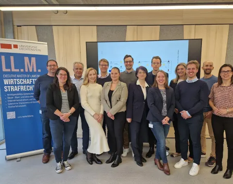 Gruppenfoto von Studierenden und Dozenten des Executive Masters of Law im Wirtschaftsstrafrecht an der Universität Liechtenstein. Im Hintergrund ein Bildschirm mit Präsentationsfolie und ein Roll-up mit Informationen zum Studiengang.