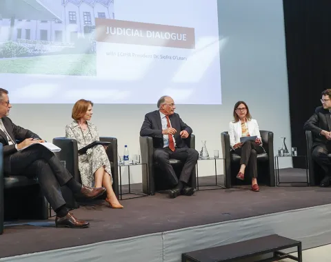 Podiumsdiskussion an der Universität Liechtenstein mit fünf Personen auf einer Bühne, die an einem juristischen Dialog teilnehmen. Im Hintergrund ist eine Präsentation mit dem Titel "Judicial Dialogue" zu sehen.