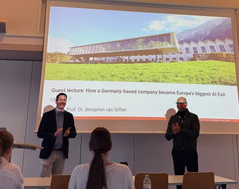 Zwei Männer stehen vor einer Präsentationsfolie in einem Vorlesungssaal der Universität Liechtenstein. Die Folie zeigt den Titel eines Gastvortrags über den Erfolg eines deutschen KI-Unternehmens. Studierende sitzen im Publikum.
