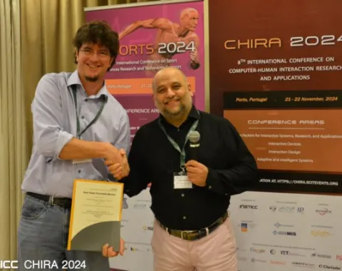 Zwei Männer stehen vor Bannern der CHIRA 2024 Konferenz. Einer hält ein Zertifikat, während der andere ein Mikrofon hält. Beide lächeln und schütteln sich die Hand. Im Hintergrund sind Details zur Konferenz in Porto, Portugal, sichtbar.