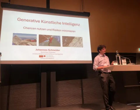 Vortragender steht vor einer Präsentation mit dem Titel "Generative Künstliche Intelligenz: Chancen nutzen und Risiken minimieren". Der Raum ist dunkel, der Fokus liegt auf der Leinwand und dem Redner.