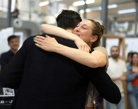 Zwei Personen umarmen sich herzlich in einem Innenraum während des LSA Sommerfests an der Universität Liechtenstein. Im Hintergrund sind unscharf weitere Personen und architektonische Details zu erkennen.