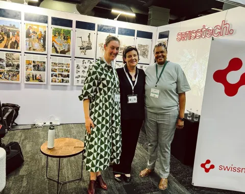 Drei Personen stehen lächelnd vor Ausstellungstafeln mit Bildern und Texten. Im Hintergrund ist ein Banner mit dem Logo von "swisstech" zu sehen. Die Szene befindet sich in einem Ausstellungsraum, der Teil der Präsentation "Making Urban Africa" auf dem South African Science Forum ist.