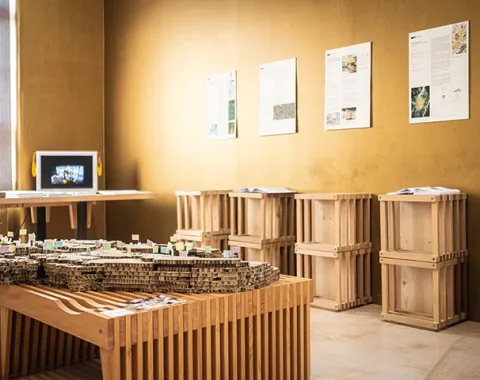 Ein Ausstellungsraum der Liechtenstein School of Architecture mit Architekturmodellen auf Holztischen und Informationsplakaten an den Wänden. Im Hintergrund ist ein Monitor zu sehen.