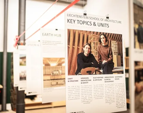 Ein Ausstellungsraum mit Architekturmodellen und Informationspostern über Lehmbau-Experten. Im Vordergrund ein detailliertes Modell eines Gebäudes mit einem roten Dach.