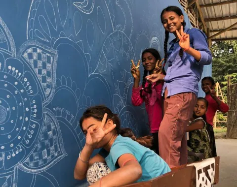 Kinder posieren fröhlich vor einer blau bemalten Wand mit kunstvollen weißen Mustern. Sie zeigen mit den Fingern das Friedenszeichen. Die Szene vermittelt Freude und Gemeinschaftsgefühl.