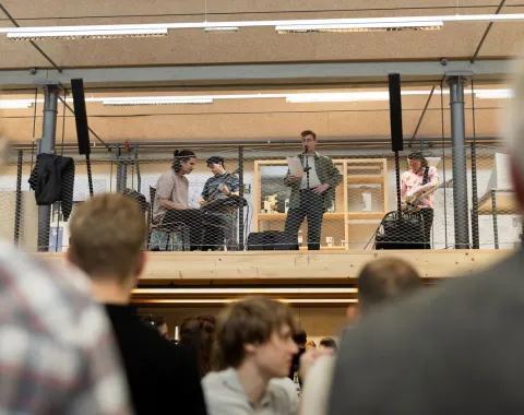 Eine Band spielt auf einer erhöhten Bühne in einem Innenraum. Im Vordergrund sind unscharf Menschen zu sehen, die dem Auftritt zuschauen. Die Szene wirkt lebhaft und ist Teil einer Veranstaltung an der Universität Liechtenstein.