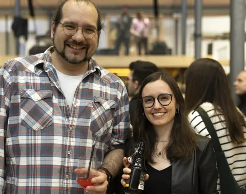 Zwei Personen lächeln in die Kamera bei einer Veranstaltung. Sie halten Getränke in den Händen. Im Hintergrund sind weitere Gäste und eine Bühne zu sehen. Die Atmosphäre wirkt fröhlich und gesellig.