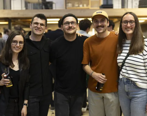 Eine Gruppe von fünf Personen lächelt in die Kamera bei einem Indoor-Event. Zwei Personen halten Getränke in der Hand. Die Umgebung ist lebhaft und gut beleuchtet, was auf eine gesellige Atmosphäre hinweist.