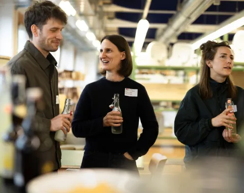 Eine Gruppe von drei Personen steht in einem Innenraum und unterhält sich fröhlich. Zwei von ihnen halten Getränkeflaschen. Im Hintergrund sind Regale und Beleuchtung zu sehen. Die Atmosphäre wirkt lebhaft und gesellig.