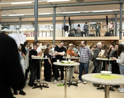 Eine Gruppe von Menschen versammelt sich in einem modernen Innenraum mit hohen Decken und großen Fenstern. Sie stehen um Stehtische, unterhalten sich und genießen die Veranstaltung. Im Hintergrund sind weitere Personen auf einer erhöhten Plattform zu sehen. Die Atmosphäre wirkt lebhaft und gesellig.