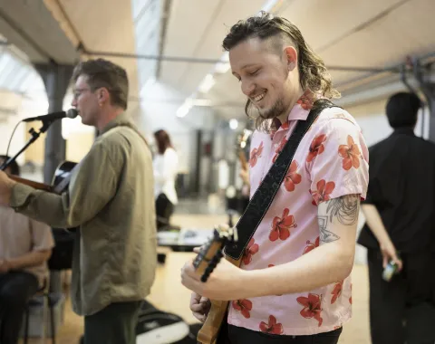 Ein Musiker in einem rosa Hemd mit Blumenmuster spielt Gitarre und lächelt. Im Hintergrund ist ein weiterer Musiker mit Mikrofon zu sehen. Die Szene findet in einem Innenraum statt, möglicherweise während einer Veranstaltung an der Universität Liechtenstein.