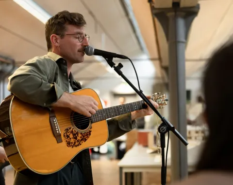 Ein Musiker spielt Gitarre und singt in ein Mikrofon bei einer Veranstaltung in einem Innenraum. Im Hintergrund sind Tische und eine helle Decke zu sehen.