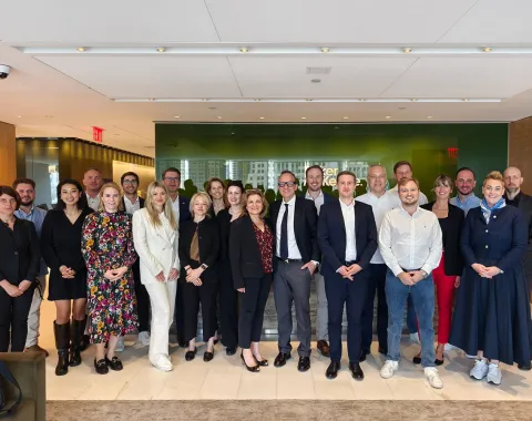 Eine Gruppe von Studierenden und Dozenten des Executive Master of Laws (LL.M.) der Universität Liechtenstein posiert in einem modernen Konferenzraum während ihrer Studienreise in New York. Sie stehen in formeller Kleidung vor einer Glaswand, die das Logo einer Anwaltskanzlei zeigt.