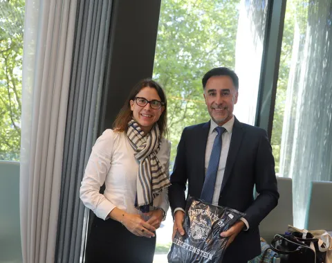 Zwei Personen stehen lächelnd in einem modernen Raum mit großen Fenstern. Eine Person hält eine Tasche mit dem Logo der Universität Liechtenstein. Im Hintergrund sind Bäume durch die Fenster sichtbar.